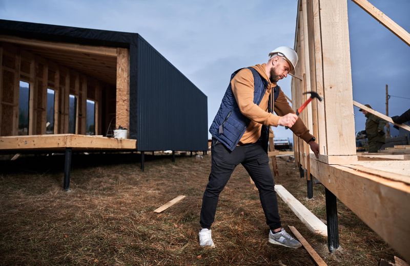 Строитель в каске и жилете забивает гвоздь в деревянную конструкцию.